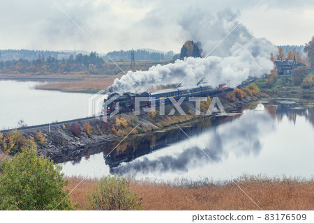 Retro steam train moves along the lake at autumn cloudy time. Karelia. Retro steam train moves along the lake at autumn cloudy time. Karelia. 83176509