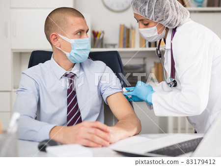 Doctor in white coat and protective mask giving vaccine injection to manager 83179412