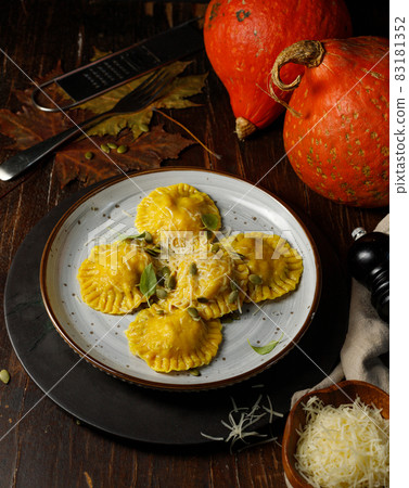 Pumpkin ravioli with ricotta and parmesan. Low key, on a dark wooden background, vertical, with a small orange pumpkin in the background Pumpkin ravioli with ricotta and parmesan. Low key, on a dark wooden background, vertical, with a small orange pumpkin in the background 83181352