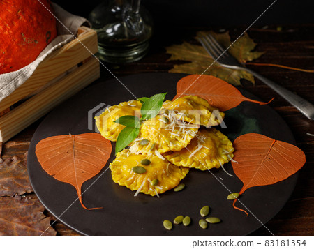 Pumpkin ravioli with ricotta on a black plate, with grated parmesan and seeds. Decorated with orange autumn leaves. Close-up 83181354
