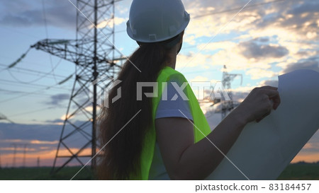 A woman power engineer with a project of new electrical works, an electrician walks against the background of high towers of power plants, the expansion of electrical equipment of voltage volts A woman power engineer with a project of new electrical works, an electrician walks against the background of high towers of power plants, the expansion of electrical equipment of voltage volts 83184457