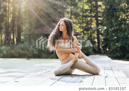 Attractive woman performs vamadevasana exercise, sage pose, sitting on a mat on a warm summer morning on a wooden bridge in the park 83185197