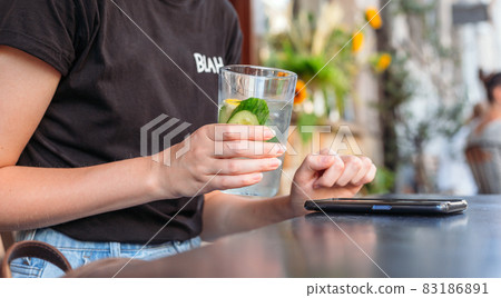 Woman at cafe drinking water and using mobile phone. Hand holding lemonade on cafe background. Glass of Cold drinks. Street front view, girl waiting for her order 83186891