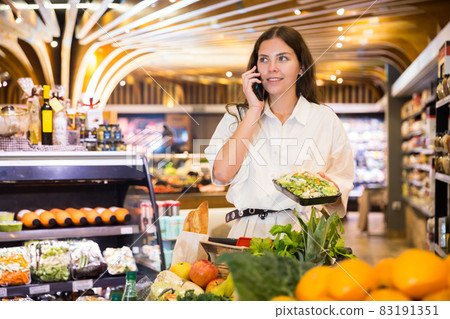Confident girl in a supermarket is talking on a mobile phone, choosing frozen vegetables 83191351