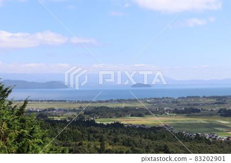 A landscape with green trees, mountains, forests, colorful fields and towns, Lake Biwa, Oki Island, a peninsula, a hazy opposite bank, mountains, clouds, and a blue sky in the foreground. 83202901