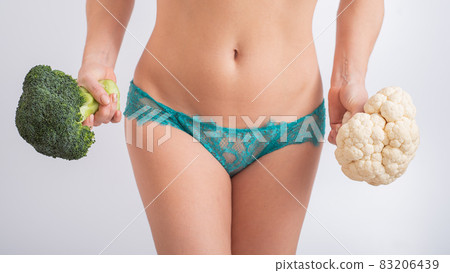 A faceless woman in panties holds cauliflower and broccoli on a white background. Food habits. 83206439