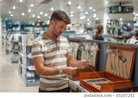 Male client in casual clothes examining and selecting fork while standing near shelf in spacious contemporary mall 83207281