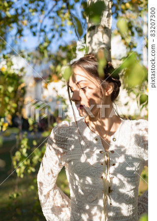Beautiful brunette woman with braids around head in a stylish vintage white lace long sleeve blouse under the birch tree. Summer time, or warm fall season 83207300