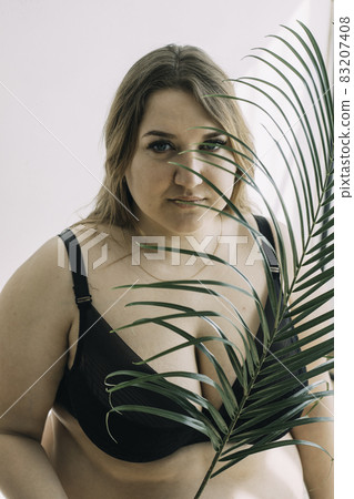 Attractive plus size girl sitting in black lingerie with a palm tree branch in her hands Attractive plus size girl sitting in black lingerie with a palm tree branch in her hands 83207408