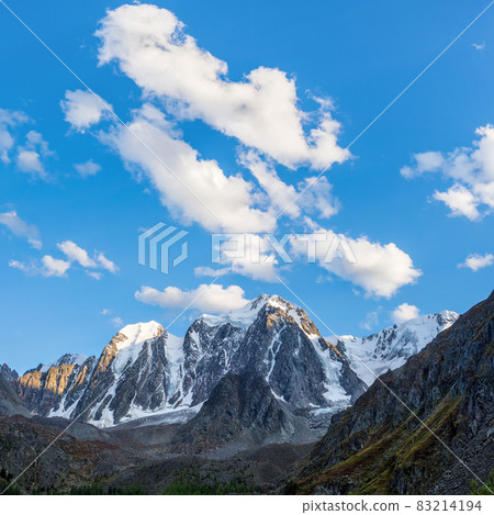 White clouds over a high pointed mountain. Snowy high-altitude alpine landscape with snow-capped mountain peak and sharp rocks under cloudy blue sky. 83214194