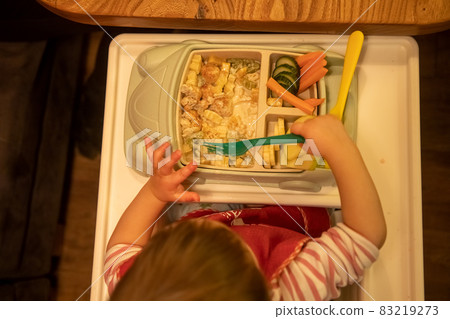 top view little cute child sits in a highchair with a plate of delicious delicious food on a table and eats with a baby fork 83219273