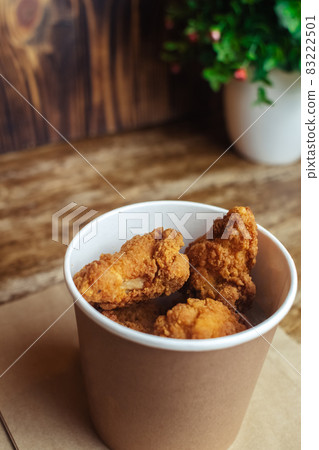 Close-up of chicken wings fried in batter. In a fast food restaurant, there is a cardboard box of chicken and sauce on the table. Sun rays fall beautifully on the hand of the girl who holds the food 83222501