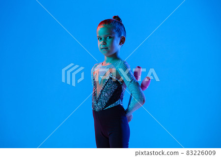 Portrait of little girl, professional rhythmic gymnast performing with gymnastic clubs isolated over blue background in neon Portrait of little girl, professional rhythmic gymnast performing with gymnastic clubs isolated over blue background in neon 83226009