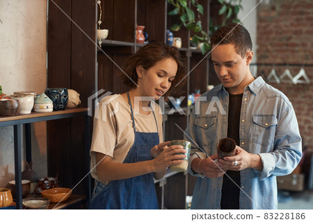 Positive young saleswoman in apron standing against shelves and showing ceramic mug to buyer in pottery store 83228186