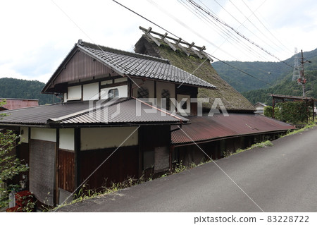 A landscape of a private house with a thatched roof, electric wires, utility poles, steel towers, mountains, and a cloudy sky 83228722
