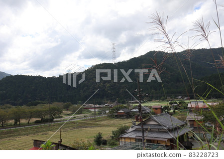 A rural landscape of thin, private houses, fields, roads, trees, bridges, rivers, mountains, utility poles, electric wires, steel towers, and clouds in the sky as seen from a hill. 83228723