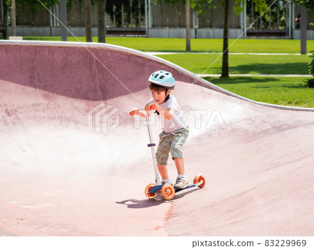 Little boy rides kick scooter in skate park. Special concrete bowl structures in urban park. Training to skate at summer. 83229969