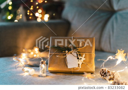 Christmas natural eco gift boxes with empty label tag. Glowing light bokeh, coniferous branch. Winter holidays mock up on gray couch background at cozy home. Zero waste, plastic free. Indoors plants. Christmas natural eco gift boxes with empty label tag. Glowing light bokeh, coniferous branch. Winter holidays mock up on gray couch background at cozy home. Zero waste, plastic free. Indoors plants. 83233082