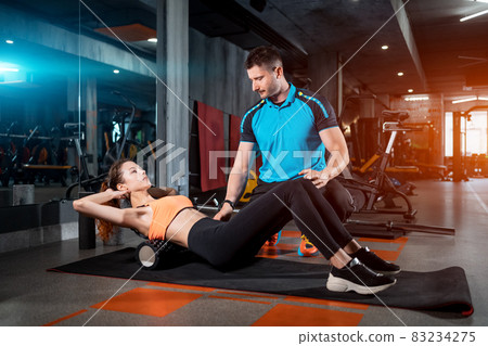 young female relaxing with myofascial release roller with trainer in gym 83234275