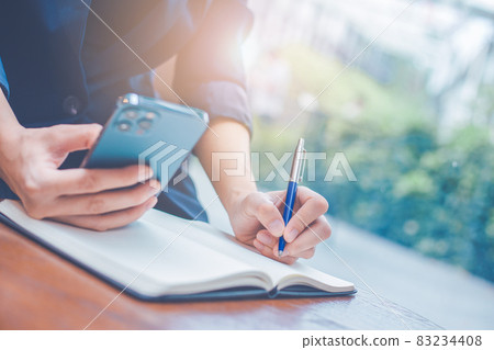 Business woman hand writing on a notepad with a pen and she is holding a cell phone in the office. Business woman hand writing on a notepad with a pen and she is holding a cell phone in the office. 83234408