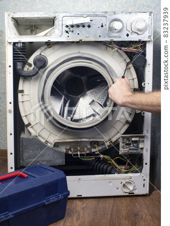 Washing machine repair. The front wall has been removed from the washing machine, the internal units and parts are visible. There is a box with a tool nearby. Repair of large household appliances 83237939