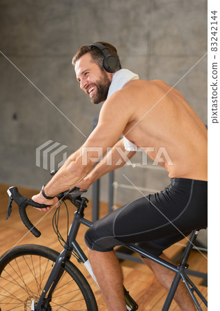 Joyful young man listening to music and riding stationery bike Joyful young man listening to music and riding stationery bike 83242144