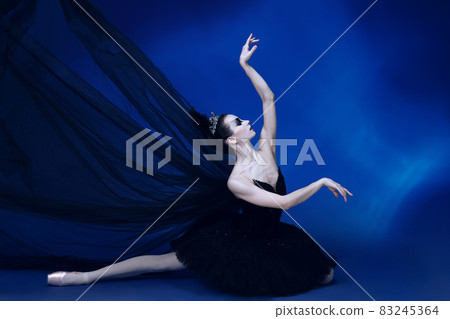 Portrait of adorable female ballet dancer in black tutu and stage make-up sitting on floor isolated on blue studio backgorund 83245364