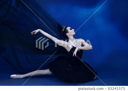 Portrait of adorable female ballet dancer in black tutu and stage make-up sitting on floor isolated on blue studio backgorund 83245373