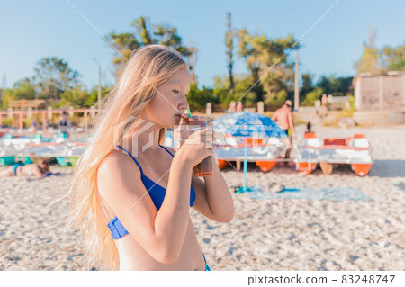 A young girl with blonde hair of European appearance, a teenager holds and drink a colored cold non-alcoholic cocktail in her hand against the background of the sea beach A young girl with blonde hair of European appearance, a teenager holds and drink a colored cold non-alcoholic cocktail in her hand against the background of the sea beach 83248747
