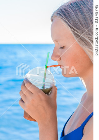 A young girl with blonde hair of European appearance, a teenager holds and drink a colored cold non-alcoholic cocktail in her hand against the background of the sea beach A young girl with blonde hair of European appearance, a teenager holds and drink a colored cold non-alcoholic cocktail in her hand against the background of the sea beach 83248748