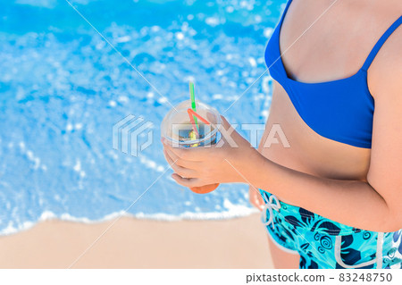 The hand of a young girl in a blue swimsuit and shorts holds a cooling, colored non-alcoholic cocktail against the background of the water of the sea beach and the shore The hand of a young girl in a blue swimsuit and shorts holds a cooling, colored non-alcoholic cocktail against the background of the water of the sea beach and the shore 83248750