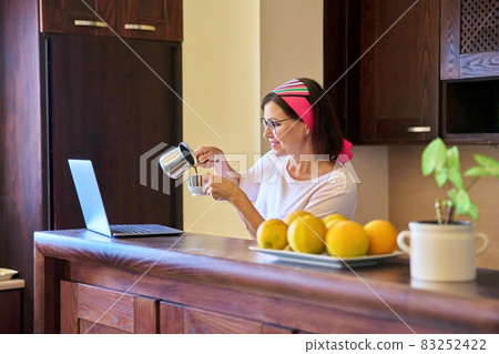 Woman in the kitchen with a cup of coffee looking into the laptop screen Woman in the kitchen with a cup of coffee looking into the laptop screen 83252422