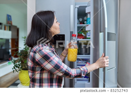 A woman opens the refrigerator at home in the kitchen, holding a bottle of orange juice. 83252743