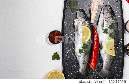 Fresh rainbow trouts on black board on table with salt, pepper, lemon and chili pepper ingredients. Tasty fishes preparing for lunch. 83254626