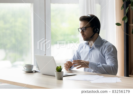 Confident man in wireless headset talking, using laptop, chatting online 83256179