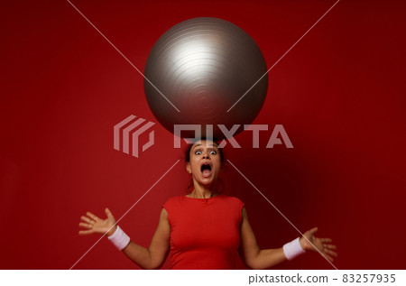 Astonished young sporty mixed race woman poses against red wall background with huge fitness ball on her head 83257935