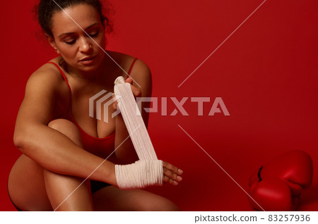 Confident Afro American female fighter, boxer sitting on a red background and tying hand wrap, preparing for the competition waving her hands with a bandage. Martial art concept with copy ad space 83257936
