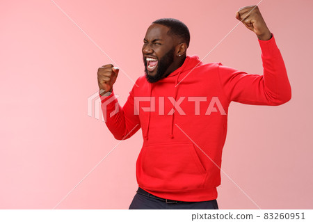 Extremely happy bearded young 25s black guy triumphing happily yelling yeah raising clenched fists celebrating success winning bet lottery standing happily pink background cheer receive great news 83260951