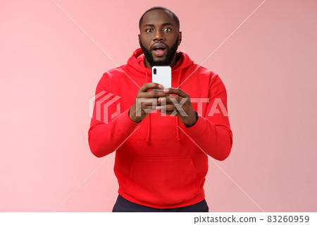 Impressed focused handsome african american bearded guy looking intrigued holding smartphone recording video capturing awesome fight, widen eyes curious surprised, standing pink background 83260959