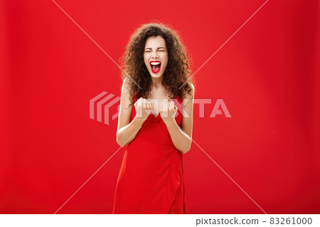 Woman triumphing and cheering joyfully raising fists near chest yelling from happiness and joy closing eyes and smiling broadly standing in red elegant dress winning lots of cash in casino 83261000