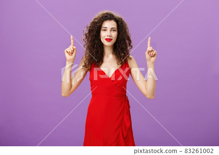 Feeling on top after making stylish makeup. Cute and tender glamourous european female in red evening dress and lipstick with dark curly hair smiling gently and pointing up with raised hands 83261002