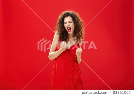 Yeah we are champions. Triumphing happy and delighted rich elegant female with curly hair in stylish dress clenching fists in yes gesture yelling celebrating joyfully win or victory over red wall Yeah we are champions. Triumphing happy and delighted rich elegant female with curly hair in stylish dress clenching fists in yes gesture yelling celebrating joyfully win or victory over red wall 83261003
