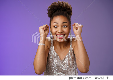 Excited african woman in elegant silver dress brilliants smiling thrilled winning clench raised fists grinning triumphing rejoicing receive perfect news celebrating, standing blue background 83261010