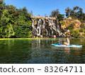 The girl swims on sup near the waterfall. Rest on the river. A group of people near an artificial waterfall. Green trees around the waterfall, blue sky with no clouds 83264711