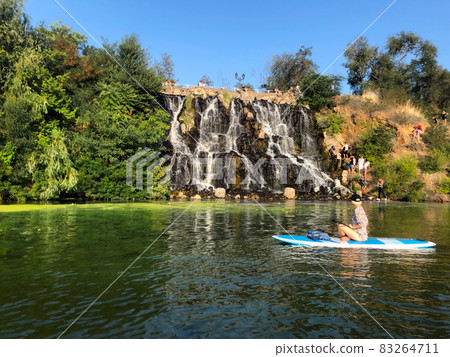 The girl swims on sup near the waterfall. Rest on the river. A group of people near an artificial waterfall. Green trees around the waterfall, blue sky with no clouds The girl swims on sup near the waterfall. Rest on the river. A group of people near an artificial waterfall. Green trees around the waterfall, blue sky with no clouds 83264711