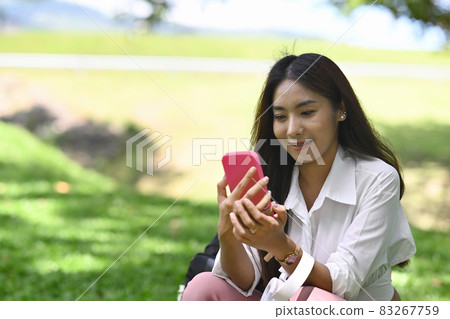 Smiling young asian woman using smart phone while sitting in the park. Smiling young asian woman using smart phone while sitting in the park. 83267759