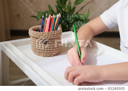 kid's hands drawing with color pencils and woven wicker basket for stationery storage. hand made basket for desktop order. crop view. kid's hands drawing with color pencils and woven wicker basket for stationery storage. hand made basket for desktop order. crop view. 83269305