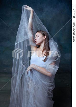Portrait of young beautiful girl, ballerina posing with white transparent cloth isolated on dark vintage studio background. 83272113