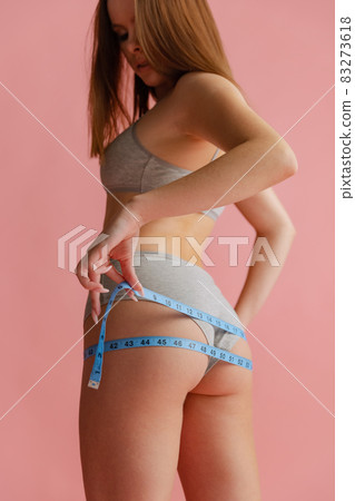 Cropped back view portrait of beautiful young woman in gray inner wear with centimeter for measuring isolated over pink background. Cropped back view portrait of beautiful young woman in gray inner wear with centimeter for measuring isolated over pink background. 83273618