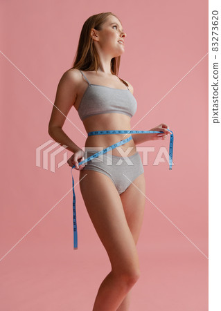 Cropped portrait of beautiful young woman in gray inner wear with centimeter for measuring isolated over pink background. Body positivity Cropped portrait of beautiful young woman in gray inner wear with centimeter for measuring isolated over pink background. Body positivity 83273620
